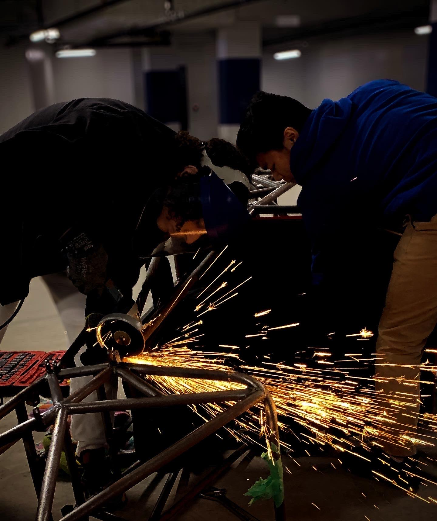 Formula SAE chassis fabrication, with team members cutting a tubular frame and sparks visible